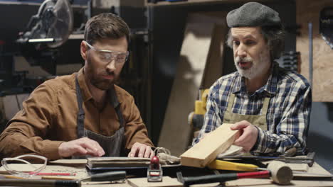 older and younger caucasian carpenters sitting at table in workshop and talking