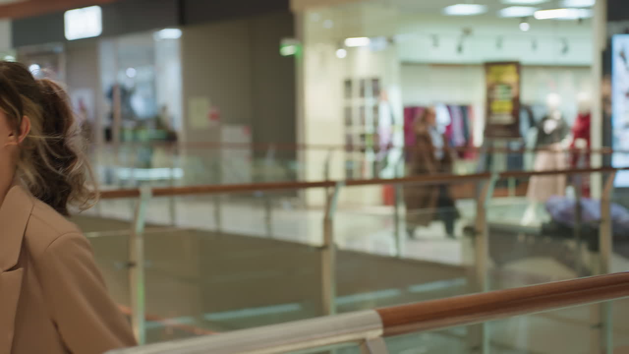 Mujer apoyada en una barandilla en una zona comercial, con escaparates a lo lejos y una señora con una bolsa amarilla descansando; mujer cansada apoyada en una barandilla rodeada de comercios y fachadas de tiendas a lo lejos.