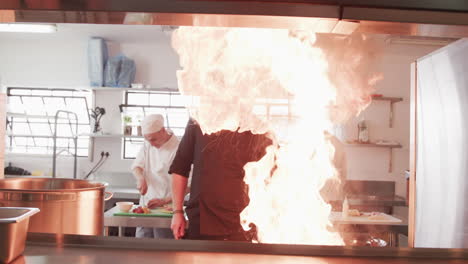 focused caucasian male chef frying food in frying pan with bursting fire in kitchen, slow motion