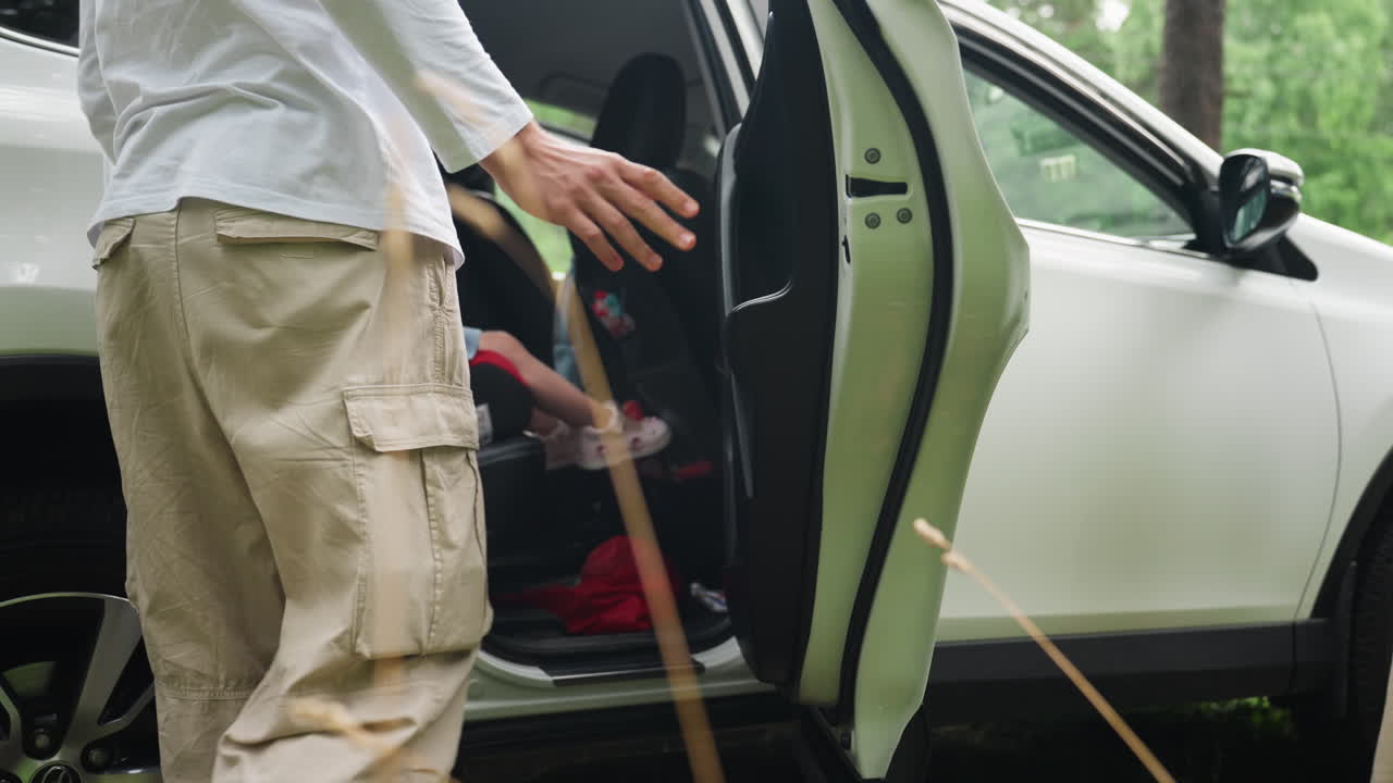 낮은 앵글에서 가족 여행 중 어린 여동생이 차 뒷좌석에 오르는 것을 부드럽게 도와주는 남자아이의 모습이 보여요. 은은한 햇빛이 따뜻한 남매의 배려심을 강조하고, 부드러움이 가득한 안전한 여행의 순간을 보여줍니다.