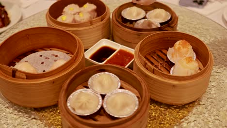 a hand uses chopsticks to pick dumplings from bamboo steamers on a restaurant table with sauces