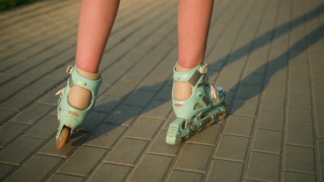 back view of person roller skating on interlocked brick path, wearing teal rollerblades, with long shadows cast on ground by afternoon sun