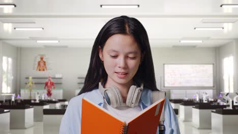 close up of asian teen girl student with a backpack holding and reading a book while standing in science laboratory