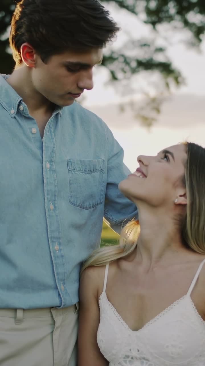 Vídeo vertical: homem sorrindo de jeans, mulher de vestido branco se inclinando ao pôr do sol no parque, luz do sol no cabelo