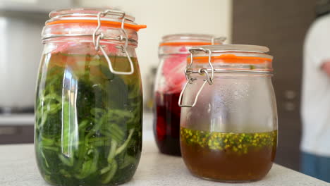 handheld shot of fermented wild garlic, fermented red berries, pickled wild garlic flowers and raspberry vinegar in jars in a kitchen with the chef working in the kitchen in the blurred background