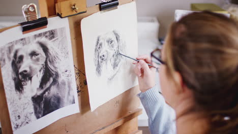 Female-Teenage-Artist-Sitting-At-Easel-Drawing-Picture-Of-Dog-From-Photograph-In-Charcoal