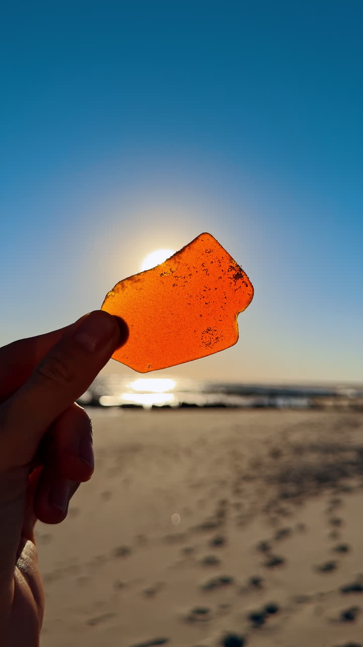 Sosteniendo un trozo de cristal naranja en la playa bajo la luz del sol. Alguien sostiene un pequeño trozo de cristal naranja mientras está de pie en la playa cerca del océano durante la puesta de sol.