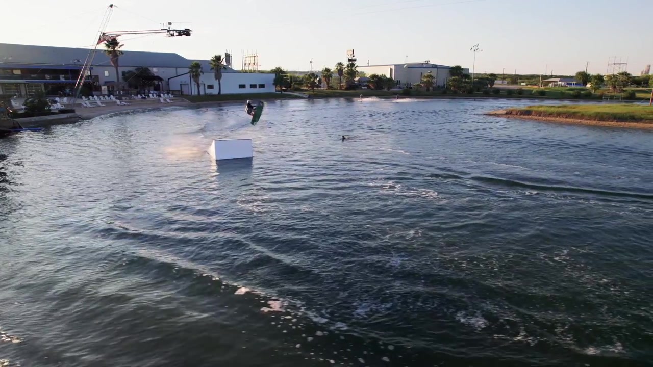 Dall'alto, un wakeboarder che esegue salti al cable park, realizzando atterraggi fluidi