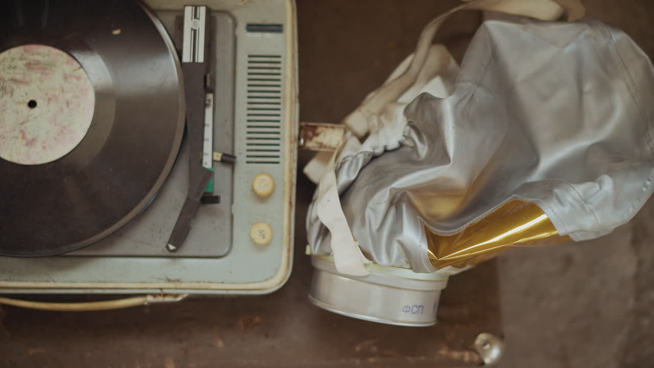Top view of dusty vintage turntable with scratched vinyl record beside crumpled silver hazmat helmet featuring reflective yellow visor, lying on weathered surface