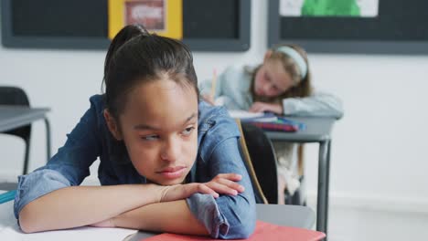 video of bored, tired biracial schoolgirl sitting at desk, leaning head on arms in class, copy space