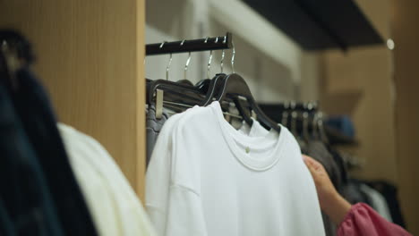 mano con tela rosa alcanza la parte superior blanca del estante de ropa, inspeccionando la textura en una tienda bien iluminada, la retira del estante, el fondo borroso muestra ropa ordenadamente dispuesta en los estantes