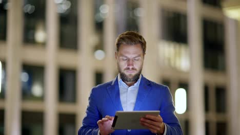 Medium shot of handsome bearded middle aged man in blue suit walking down evening street and browsing net on tablet pc