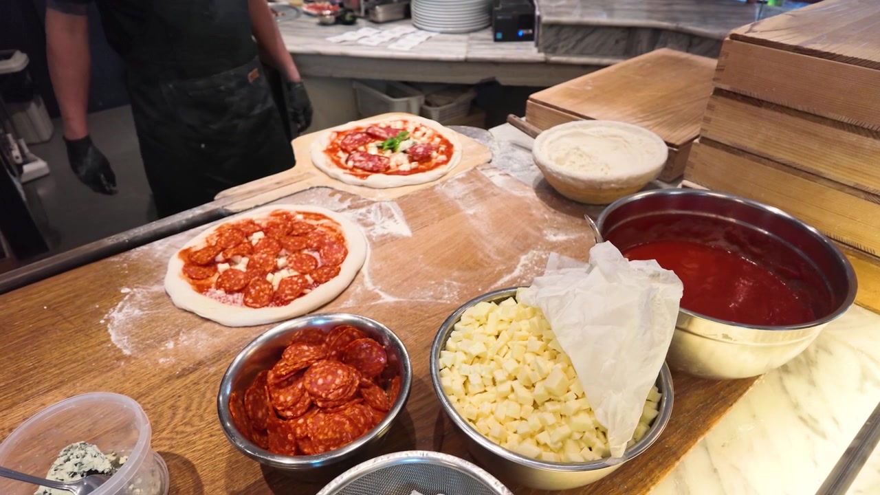 Koch belegt Peperoni-Pizza auf Holzarbeitsplatte mit frischen Zutaten unter heller Küchenbeleuchtung