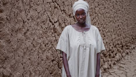 young african woman wearing traditional clothing standing against mud wall, embodying cultural heritage and resilience with direct gaze and natural surroundings