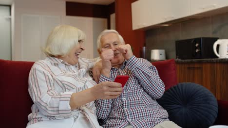 Glückliches-Lächelndes-älteres-Paar-Mit-Mobiltelefon-Zu-Hause.-Ausruhen-Auf-Dem-Sofa-Im-Gemütlichen-Wohnzimmer