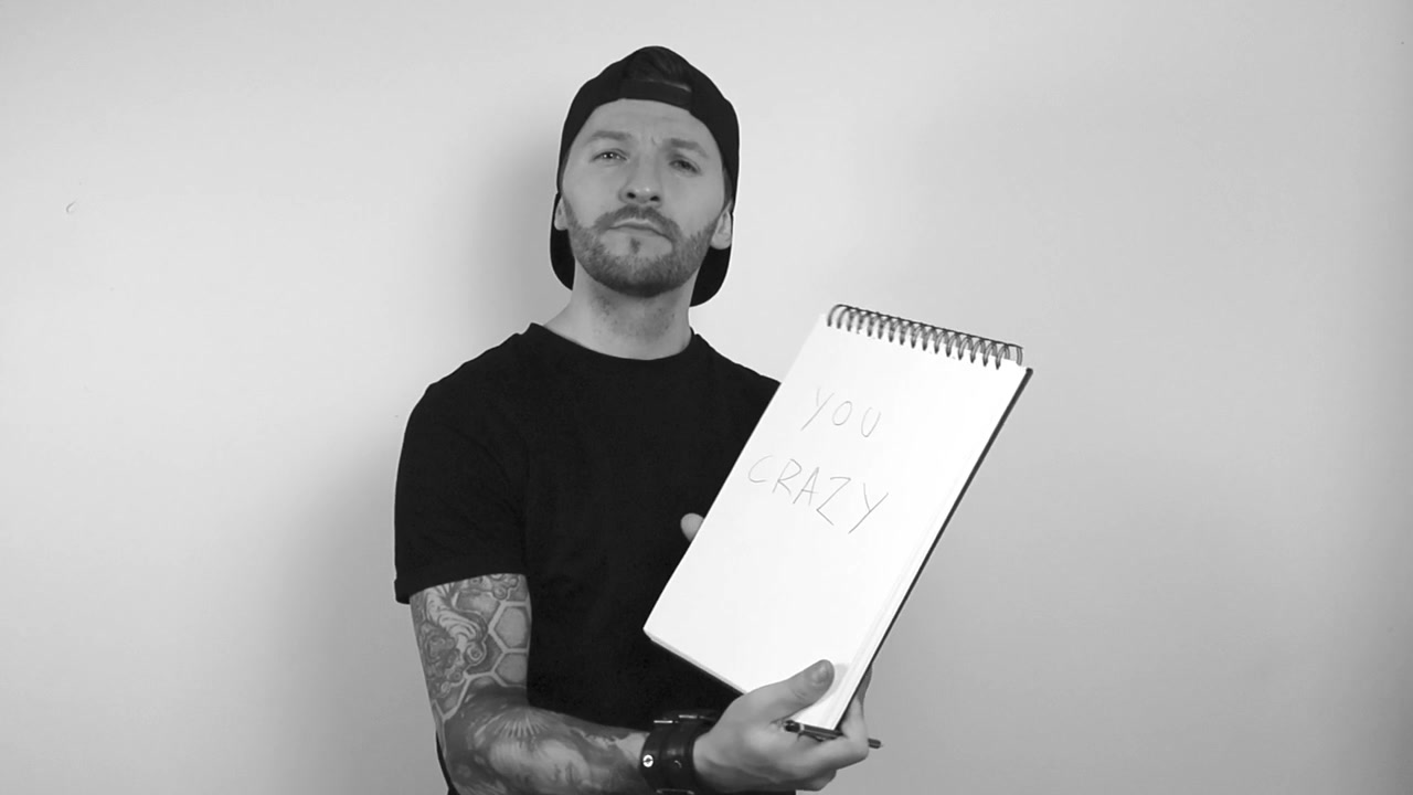 Gråtoneopptak av en tatovert mann som lytter til noen som uttrykker bekymringer og skriver ned ting i notatboken sin mens han nikker og tenker over det. Mannen viser notatbokskrift som sier Du er gal