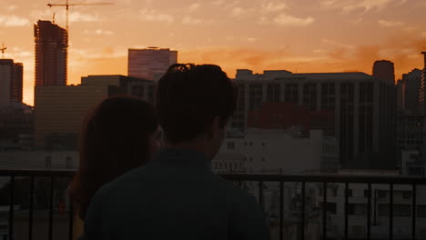 rear view of couple on rooftop terrace looking out over city skyline at sunset