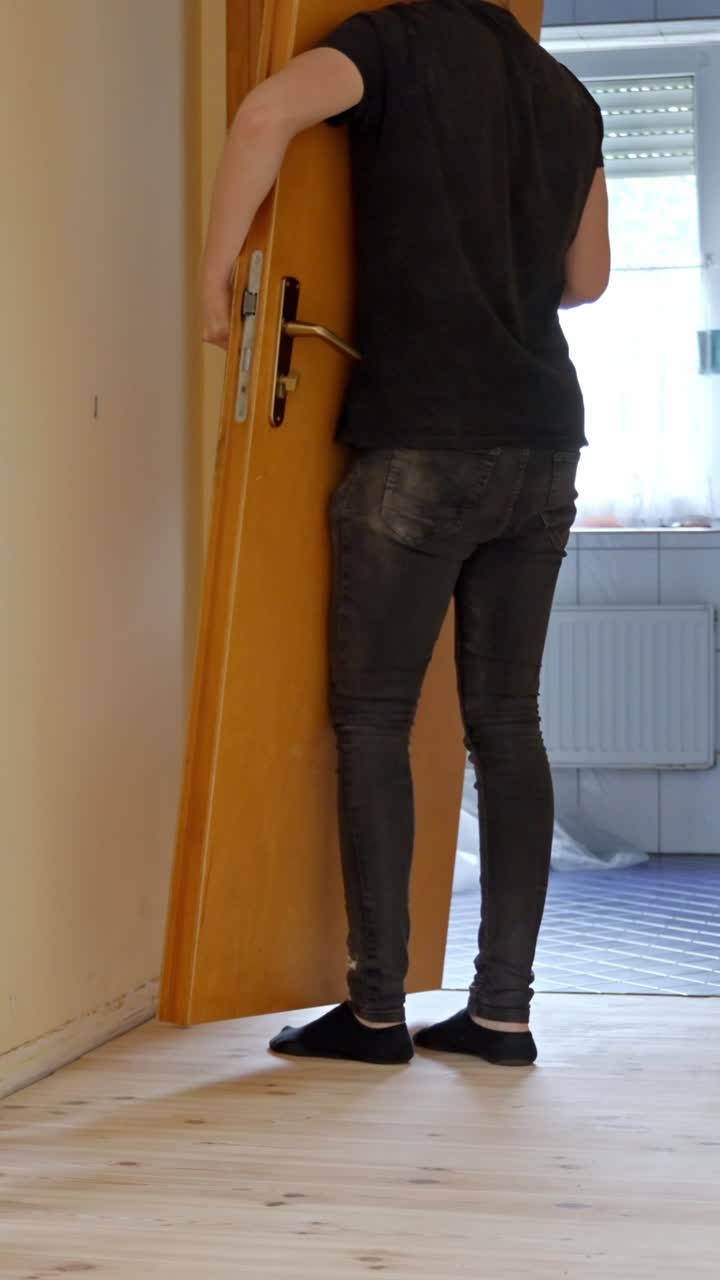 A man in casual clothes removes a wooden door from its hinges during a home renovation, standing on freshly sanded light wood flooring in a room lit by daylight, vertical orientation