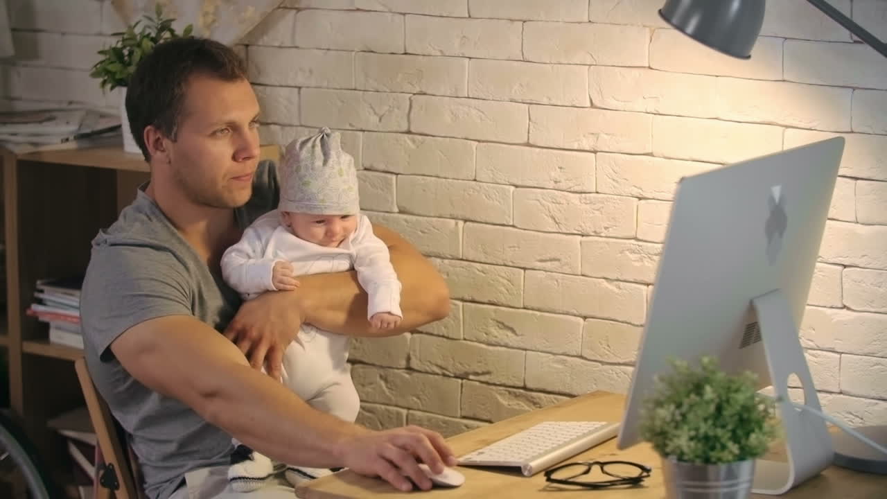 Young Father Holding His Baby While Working On The Computer At Home