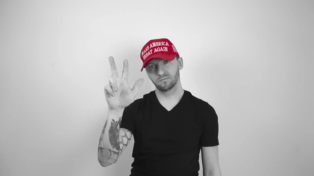 Grayscale footage of a man with tattoos, short hair and a beard who is wearing a red MAGA Make America Great Again hat counting to three with his fingers and then pointing and looking at the camera.