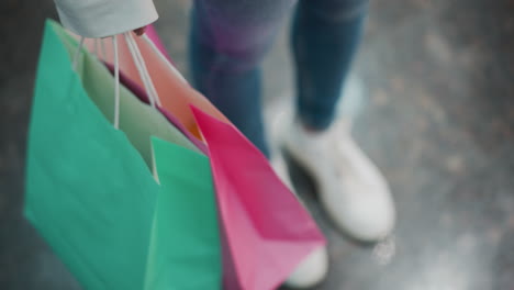 close-up of person holding green and pink shopping bags while standing on shiny marble floor, blurred white sneakers in background enhance modern retail vibe with subtle motion of swaying bags