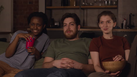 tres compañeros de cuarto están viendo una película mientras comen palomitas de maíz y papas fritas sentados en un sofá en la oscuridad en casa