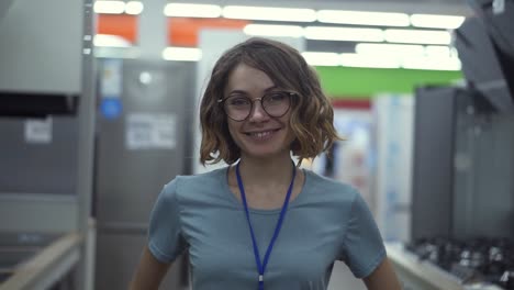 Positive-female-seller-or-shop-assistant-portrait-in-supermarket-store.-Woman-in-blue-shirt-and-badge-looking-at-the-camera-and-smiling.-Household-appliances-on-the-background