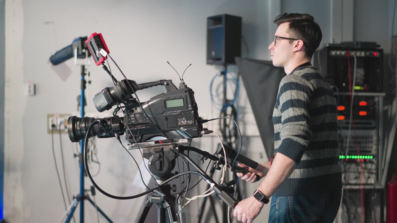 Man operating professional video camera in studio setting. Adjusting camera equipment on tripod during filming. Technician using camera rig with additional accessories in production studio