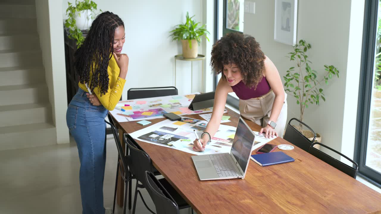 Diversas compañeras que colaboran en el diseño en una oficina en casa utilizando hojas impresas y un portátil