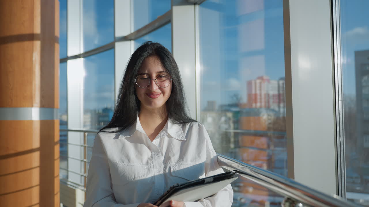 Smiling woman in white shirt holding tablet while calling out to camera with cheerful gesture, standing by large window overlooking urban cityscape on sunny day, radiating friendliness and warmth