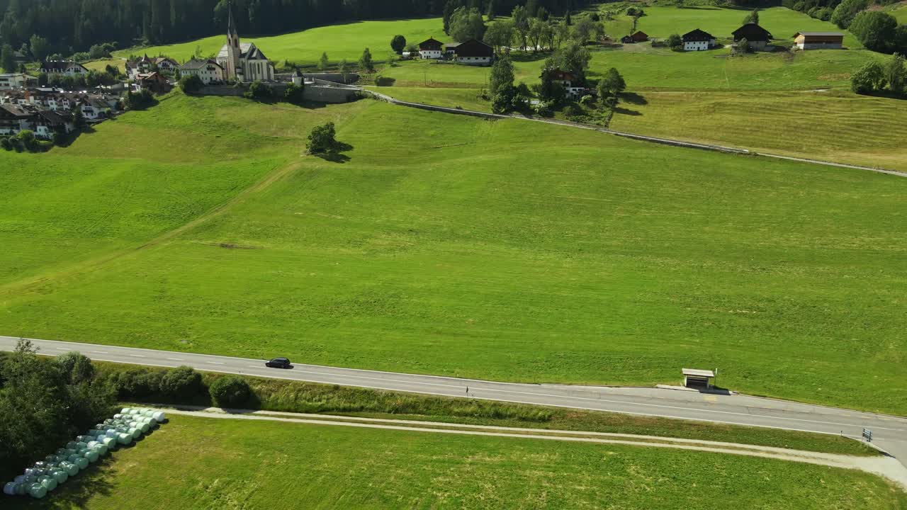 A black car drives along a rural road that cuts through lush green fields. In the background, a picturesque village with a tall church tower sits peacefully against a forested hillside.