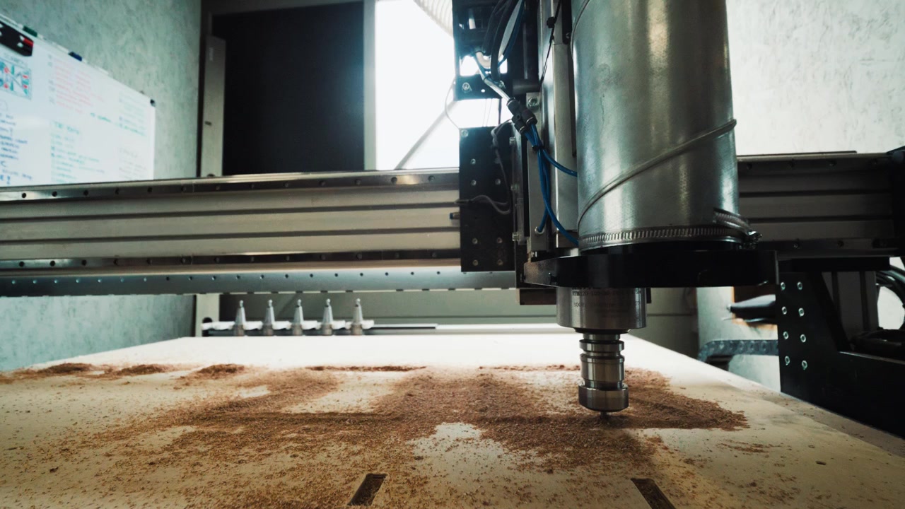 Ampia inquadratura dal basso di un router CNC su un foglio di compensato polveroso con i percorsi di taglio visibili.