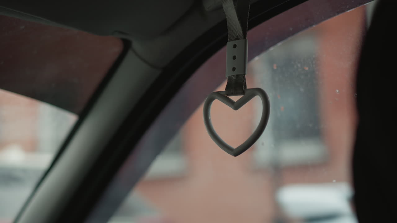 Interior passenger side view of heart shaped handle swinging inside moving car cabin against blurred urban background during winter commute showing motion and ambient atmosphere under overcast sky