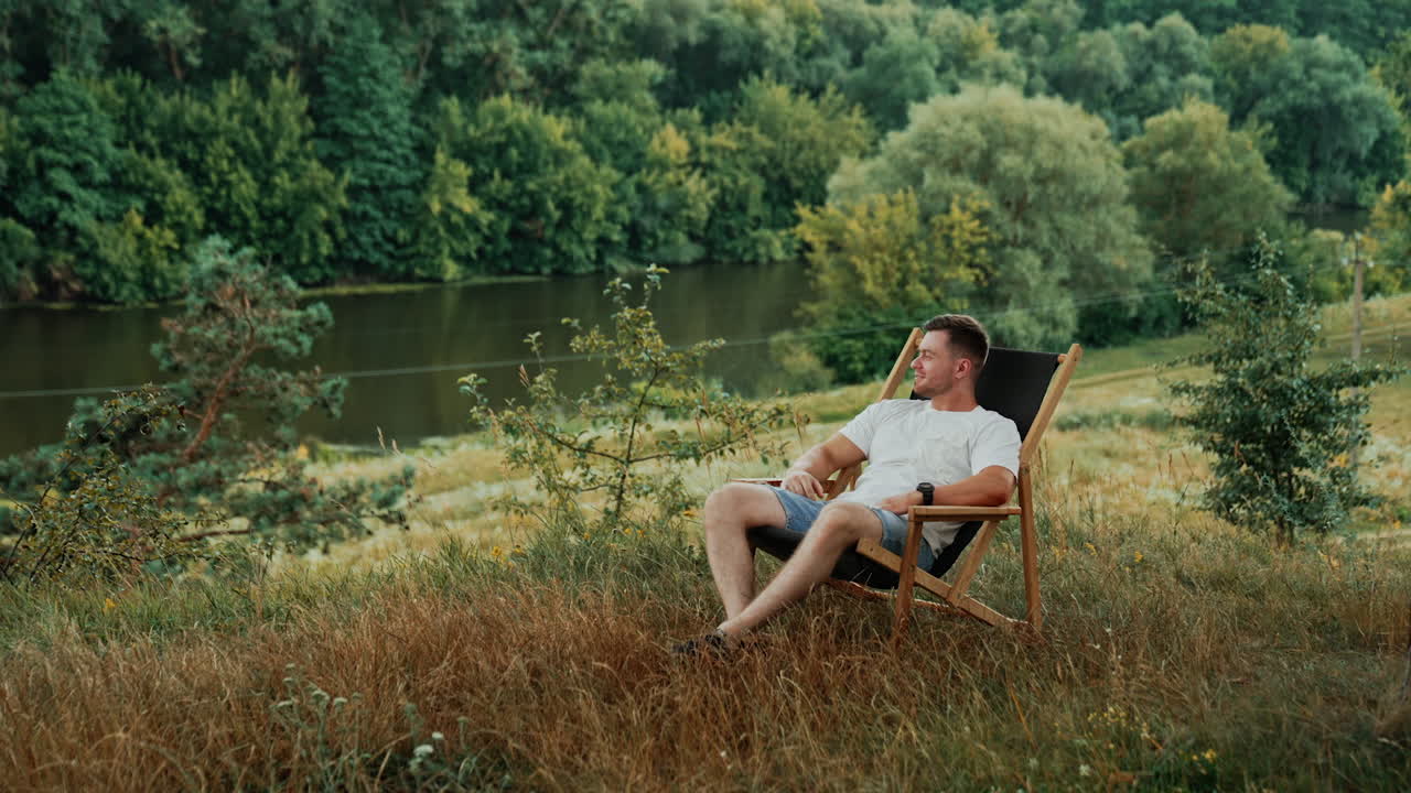 Bearded Caucasian man in white t-shirt sits in a folding chair. Happy relaxed man smiles enjoying the nature landscape.