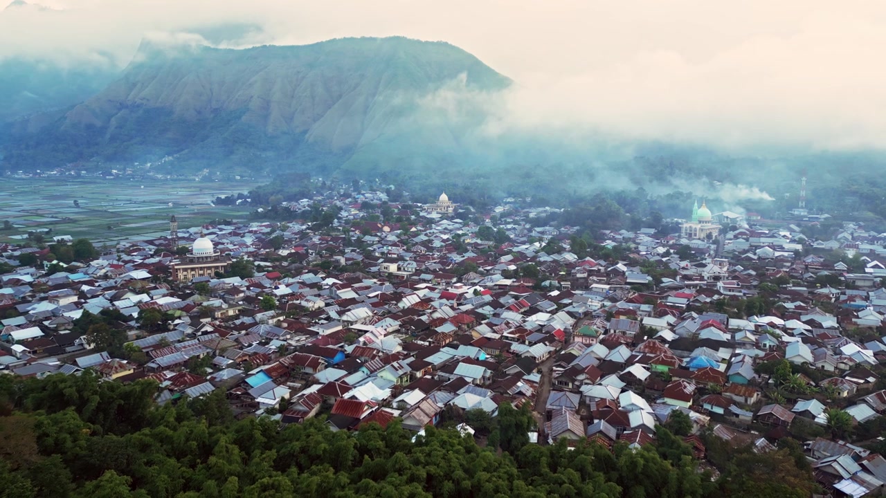 Kota pedesaan Indonesia yang dikelilingi lanskap vulkanik, awan rendah, dan hutan lebat dengan masjid serta permukiman desa, rekaman drone udara dengan gerakan maju