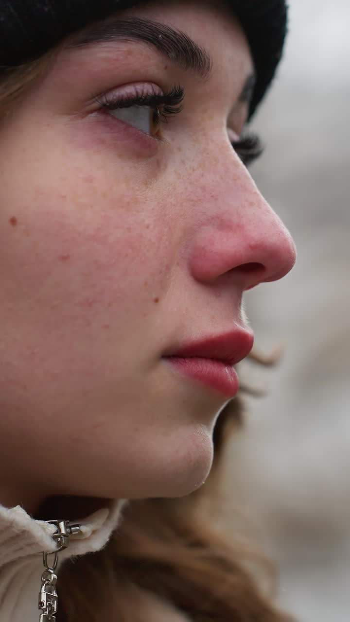 Close up side shot of young girl in black cap and brown jacket walking with serious expression and wind-blown hair against blurred city background under cold overcast sky during quiet winter day