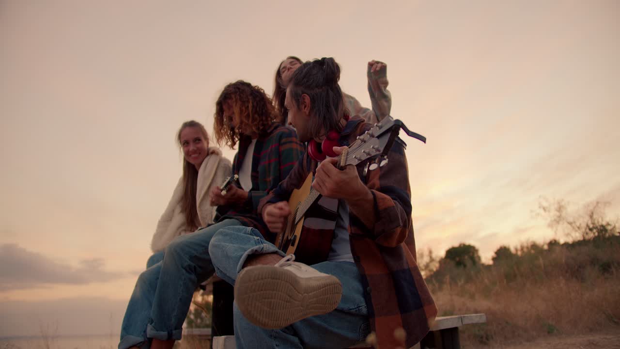 Two guys are sitting and playing guitars. Two girls listen to their boyfriends playing guitars against the background of the sunset. Rest outside the city. Rest in the country house