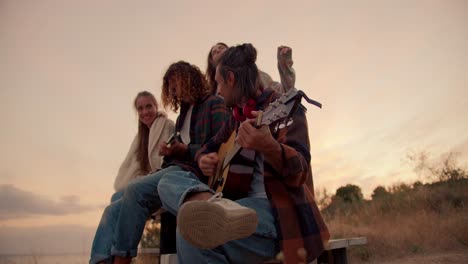 Two-guys-are-sitting-and-playing-guitars.-Two-girls-listen-to-their-boyfriends-playing-guitars-against-the-background-of-the-sunset.-Rest-outside-the-city.-Rest-in-the-country-house