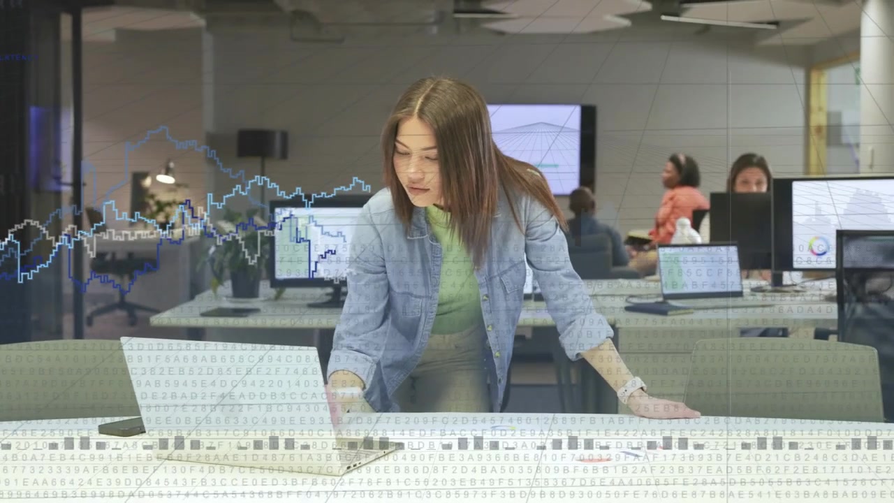 Ricevendo un aggiornamento del grafico, una donna si china sul laptop prendendo appunti mentre controlla i grafici a parete per rivedere i dati.