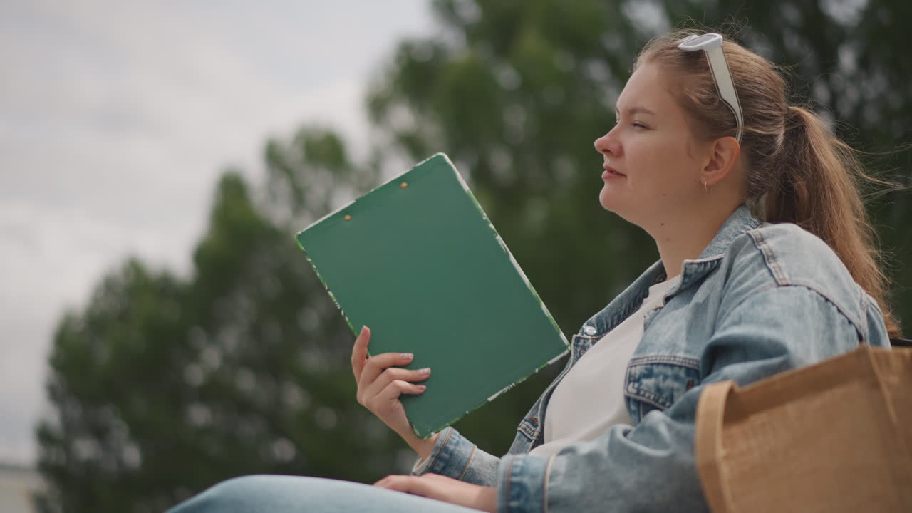 Donna bianca seduta all'aperto che tiene una cartella, giacca di jeans, borsa di tela, sfondo di un parco alberato, che guarda il cielo con espressione pensierosa, alternando lettura e appunti, le inquadrature suggeriscono.