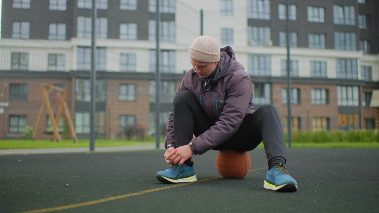 white man tying sneaker on court, wearing puffer jacket and knit beanie, sitting on basketball near hoop at residential playground, overcast sky and cool morning, focused on lacing footwear