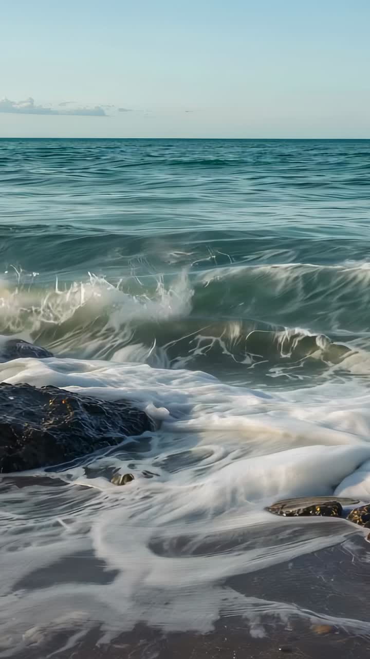 Vertical video: Approaching swell lifting waves at rocky shore, breaking on dark rocks leaving foam