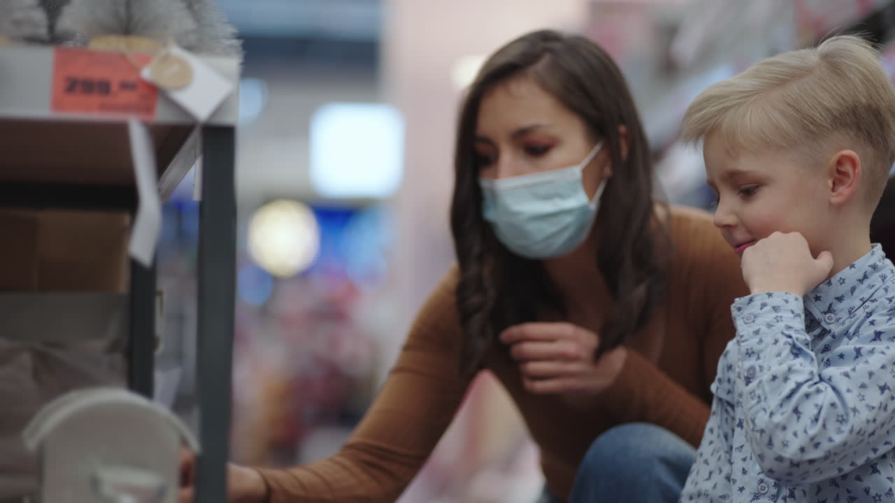 얼굴 마스크를 쓴 전염병 여성과 아들이 어린이 가게에서 장난감을 선택하고 있는 젊은 어머니가 슈퍼마켓에서 물건을 사고 있습니다. 바이러스 질병 예방