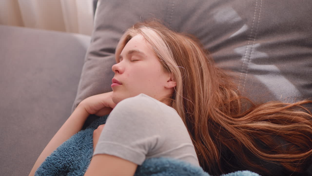 Close up of relaxed woman sleeping sideways on soft grey cushion as sunlight passes through white curtains above head while she gently adjusts position during peaceful indoor rest moment