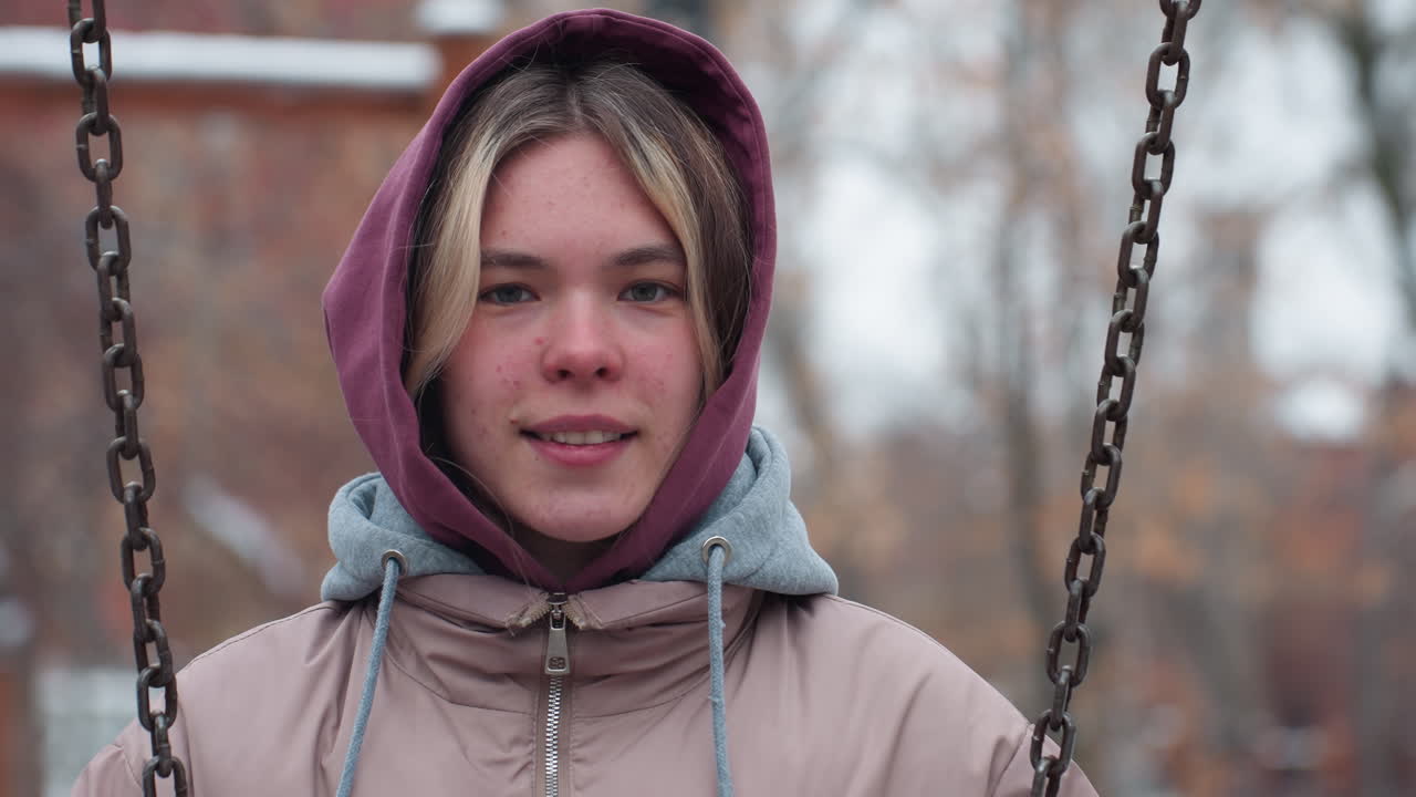 joven con traje de invierno sosteniendo una cadena de columpio, mirando a la distancia con una expresión reflexiva, la cara tiene manchas naturales, y el fondo borroso de árboles y un entorno al aire libre nevado