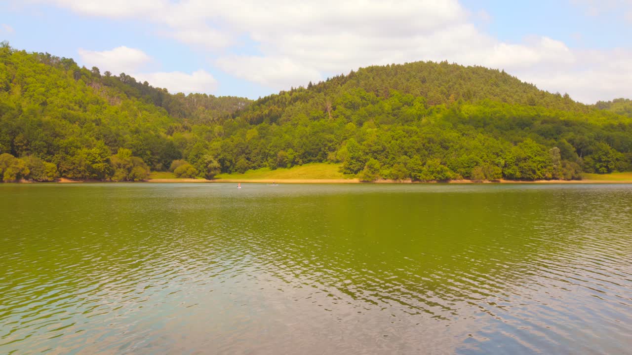 Wide still shot of Lac de Mondély in France. Calm green water and forest-covered hills under a partly cloudy summer sky