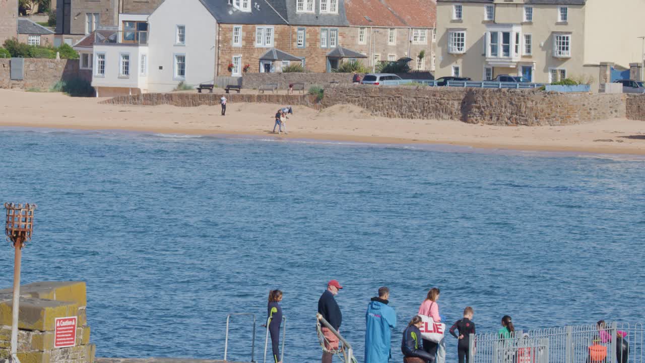 A group of people strolls along a pier by the water in a sunny coastal town, with historic stone houses, sandy beach, and calm daylight lighting
