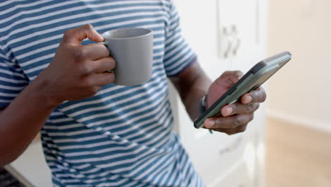 Happy-african-american-man-drinking-coffee-and-using-smartphone-in-sunny-kitchen,-slow-motion