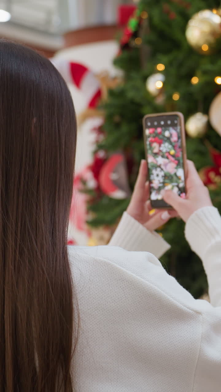 Back view of woman taking picture of tall Christmas tree adorned with ornaments and lights in a vibrant mall, bright lights filter through the mall modern architecture