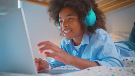 boy in bedroom lying on bed wearing wireless headphones using laptop computer